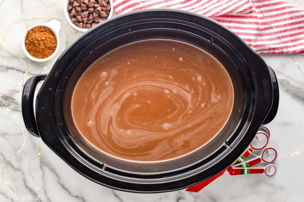 overhead of slow cooker of crockpot hot chocolate with teaspoons, cocoa powder, and chocolate chips sitting beside.