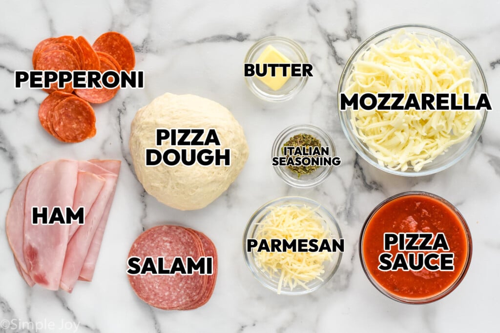 overhead of pepperoni, ham, salami, pizza dough, butter, italian seasoning, parmesan, pizza sauce, and mozzarella in bowls and labeled