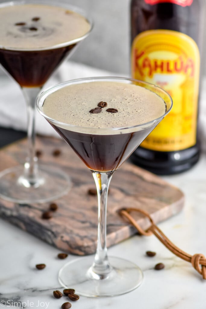 two martini glasses of espresso martinis garnished with coffee beans with coffee beans surrounding and bottle of Kahlua in the background