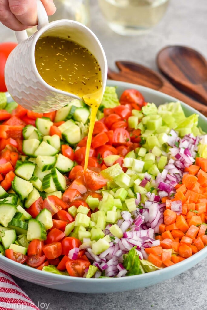 Overhead photo of a bowl of ingredients for Chopped Salad recipe with person's hand pouring salad dressing over ingredients.