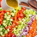 Overhead photo of a bowl of ingredients for Chopped Salad recipe with person's hand pouring salad dressing over ingredients.