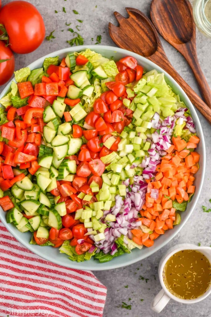 Overhead photo of a bowl of ingredients for Chopped Salad recipe. Tomatoes, salad dressing, and salad tongs are on counter beside bowl.