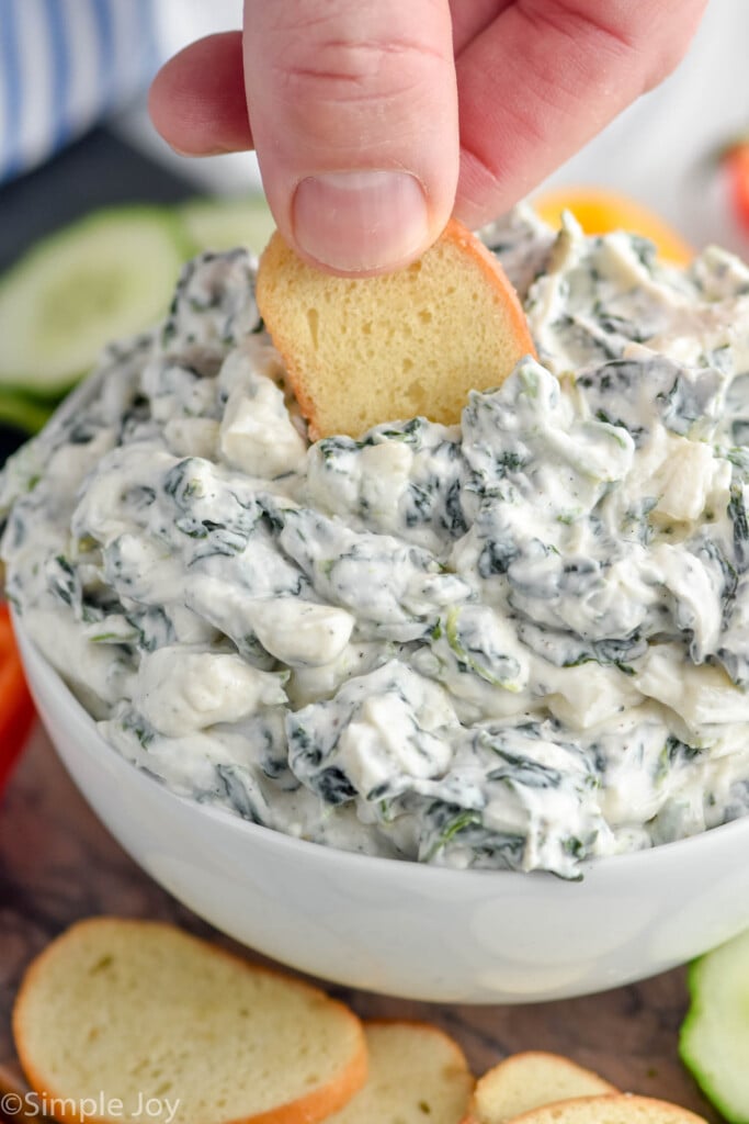 Spinach Dip Recipe served in a bowl. Sliced peppers, cucumbers, and chips beside bowl for dipping. Person's hand dipping chip into Spinach Dip.