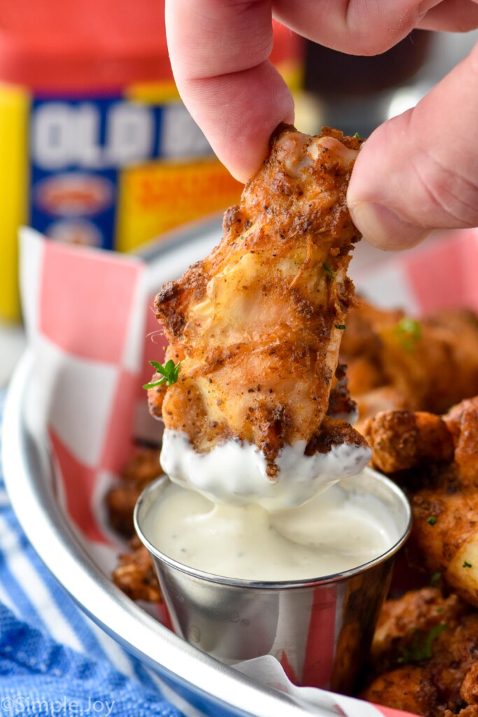 Close up photo of person's hand dipping Old Bay Chicken Wing into homemade ranch dressing. Container of Old Bay seasoning is in the background.