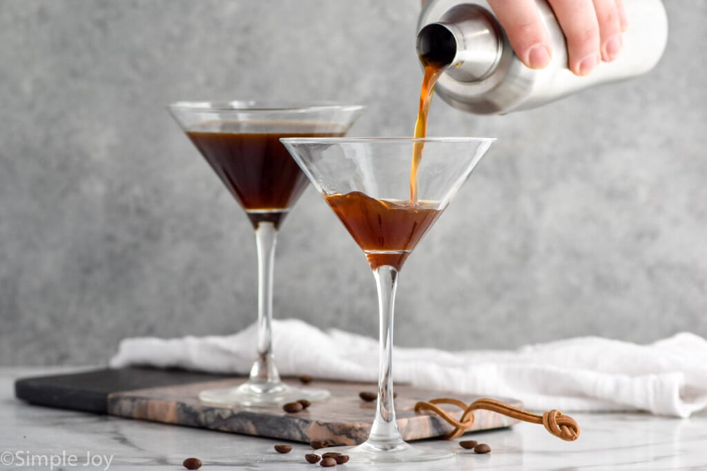 man's hand pouring cocktail shaker of espresso martini ingredients pouring into a martini glass surrounded by coffee beans and martini glass of espresso martini sitting in background