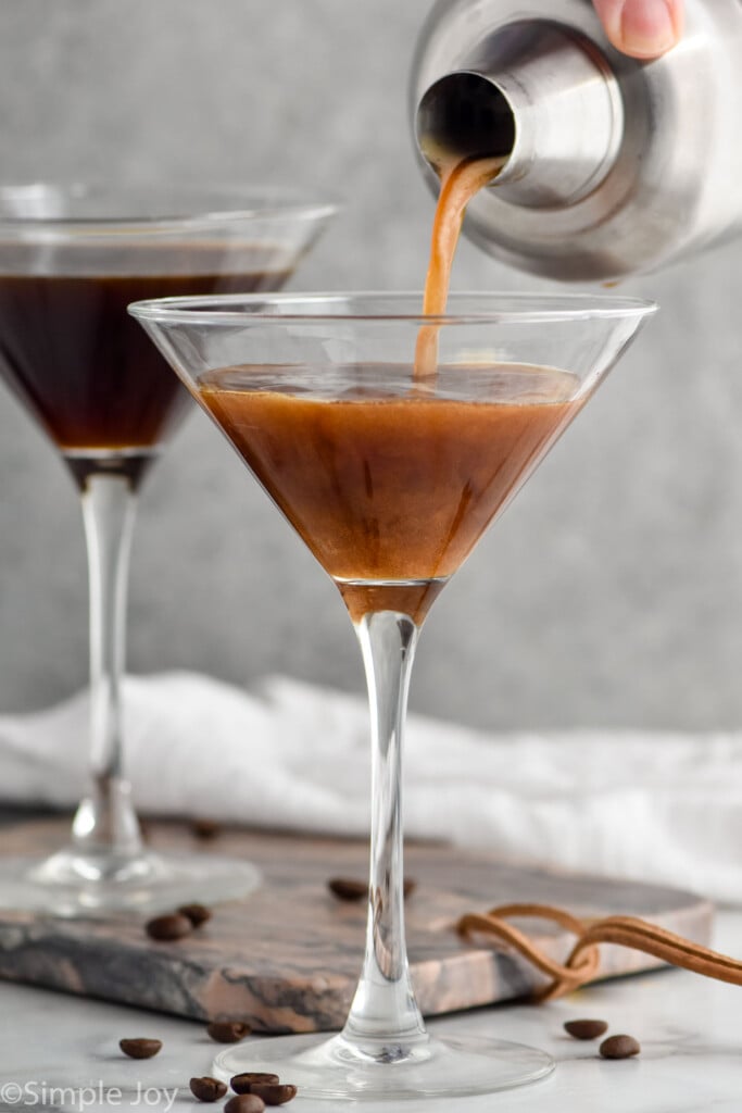 cocktail shaker of espresso martini ingredients pouring into a martini glass surrounded by coffee beans and martini glass of espresso martini sitting in background