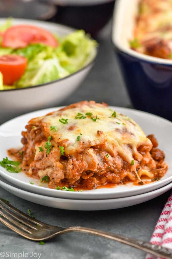 Close up photo of a piece of Homemade Lasagna served on a plate with side salad in the background.