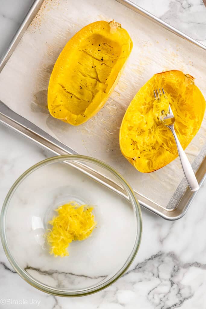 overhead of baked spaghetti squash on a rimmed baking sheet that is be scraped with a fork and some of the inside of the squash in a bowl next to the baking sheet