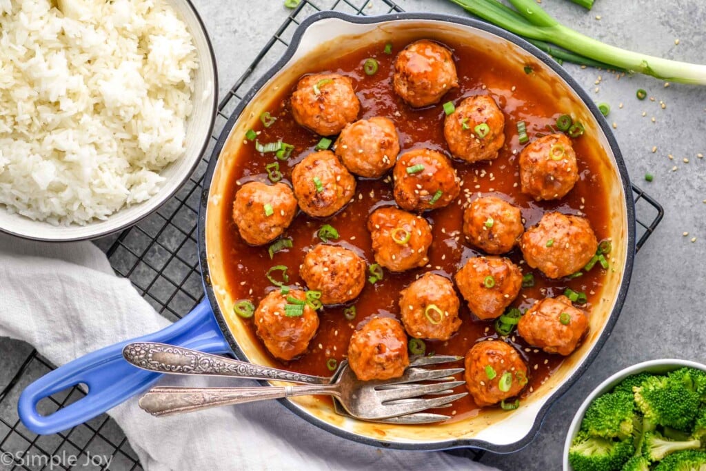 Overhead photo of Teriyaki Meatballs recipe cooking in a pan. Bowls of rice and broccoli are beside pan.