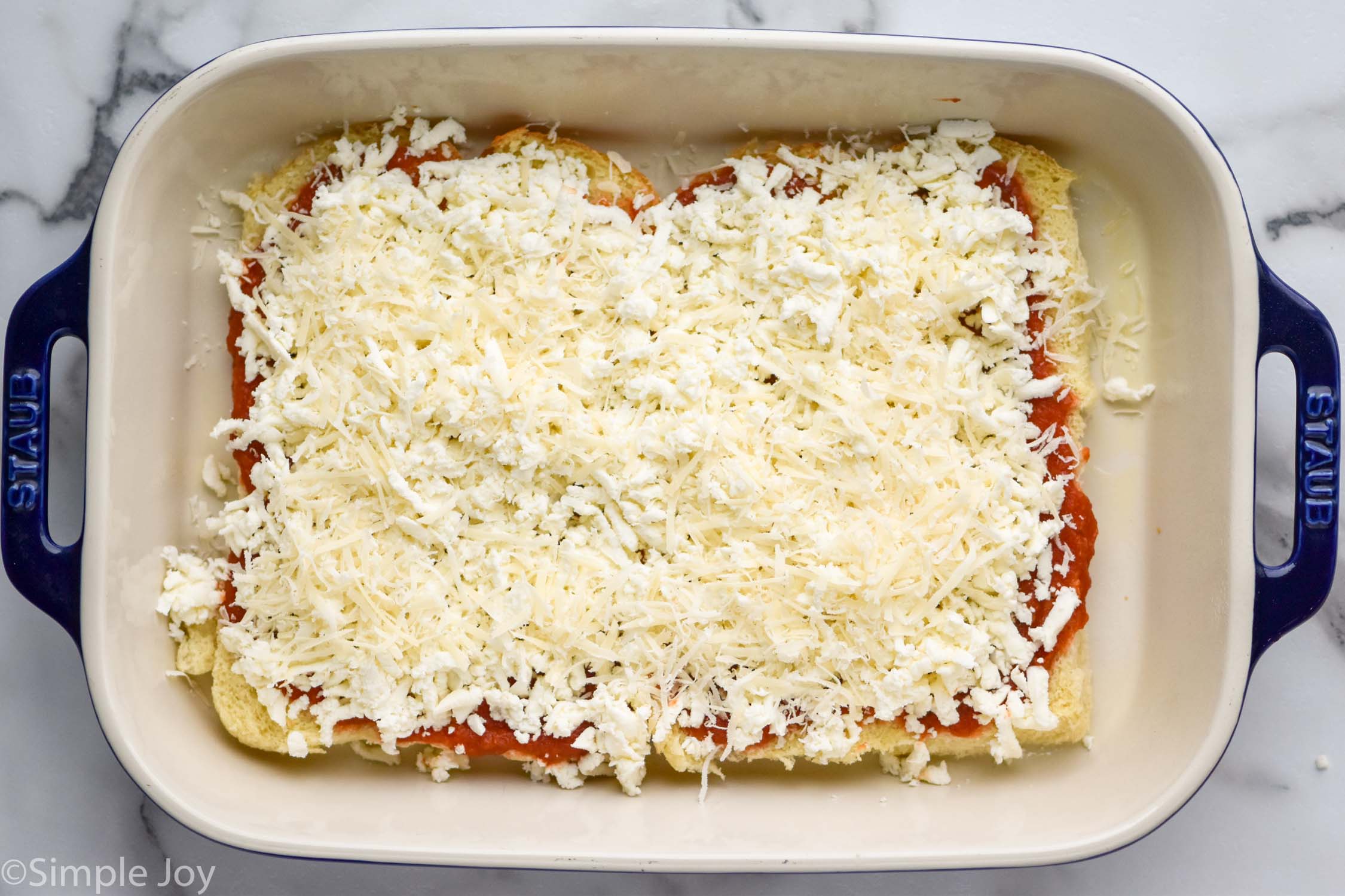 Overhead photo of baking dish of layered ingredients for Pizza Sliders recipe with cheese on top.