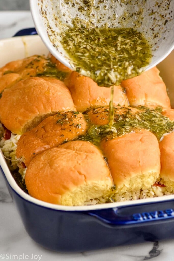 Close up photo of bowl of butter sauce being poured onto tops of rolls in baking dish for Pizza Sliders recipe.