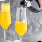 Pinterest graphic for Mimosa recipe. Text says, "the best Mimosa recipe simplejoy.com" Image is photo of person's hand pouring champagne into flute glass of orange juice for Mimosa recipe.