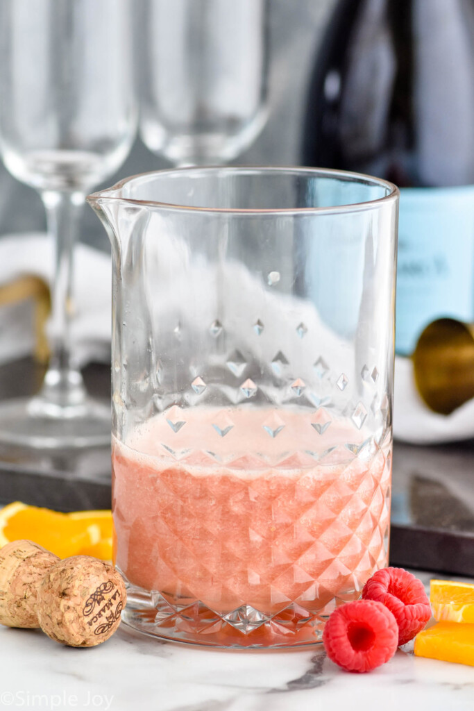 Photo of a pitcher of juice for Raspberry Mimosa recipe. Orange slices and raspberries on counter beside pitcher, and bottle of sparkling wine and flute glasses in the background.