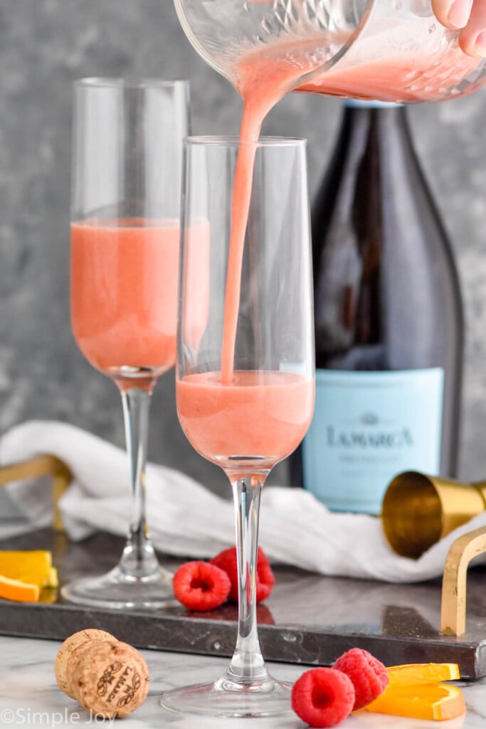 Photo of person's hand pouring juice into flutes for Raspberry Mimosa recipe. Bottle of sparkling wine in background. Raspberries and orange slices on counter beside flutes.