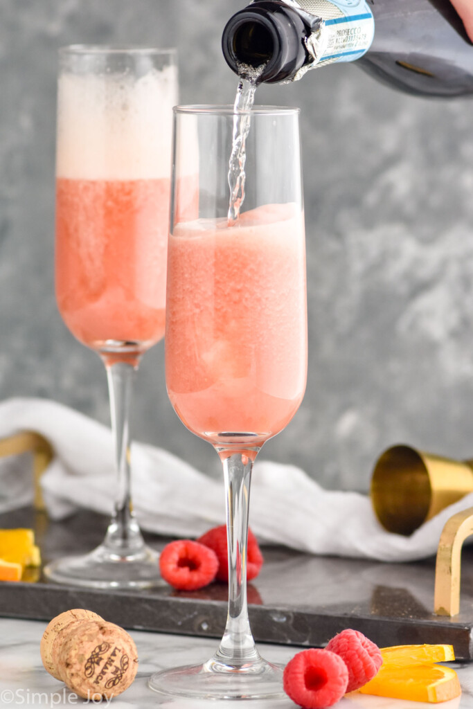 Photo of person's hand pouring sparkling wine into a flute of ingredients for Raspberry Mimosa recipe. Raspberries and orange slices on counter beside flutes.