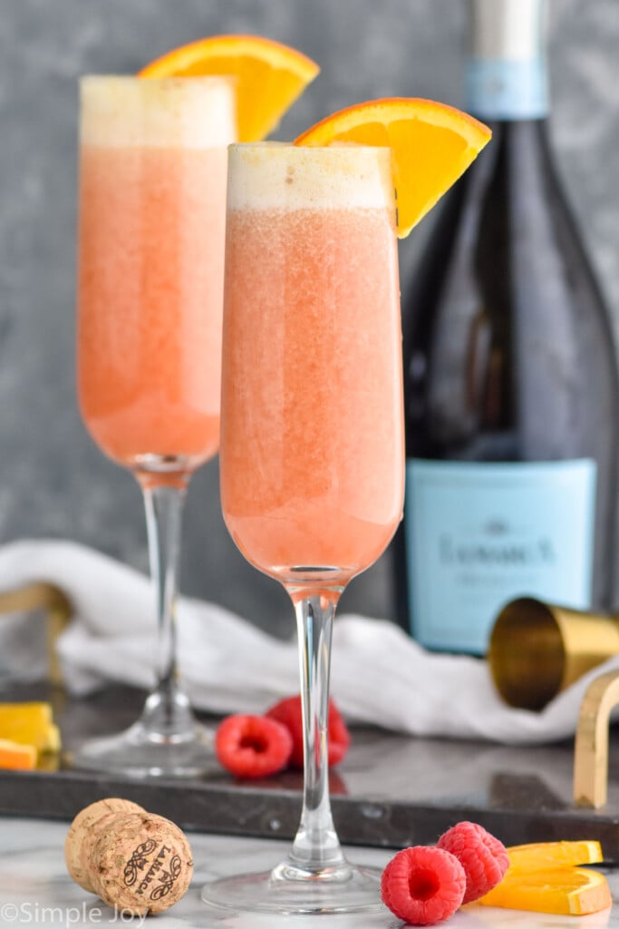 Photo of Raspberry Mimosas garnished with orange slices. Bottle of prosecco wine in background. Orange slices and raspberries on counter beside flutes.