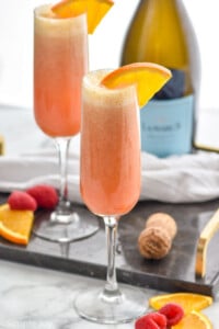 Photo of Raspberry Mimosas garnished with orange slices. Bottle of prosecco wine in background. Orange slices and raspberries on counter beside flutes.