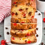 Photo of a loaf of Ambrosia Bread sliced on a cutting board. Two cups of coffee on counter, and cherries and chocolate chips on cutting board beside bread.