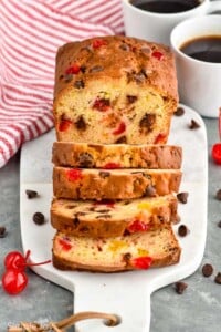 Photo of a loaf of Ambrosia Bread sliced on a cutting board. Two cups of coffee on counter, and cherries and chocolate chips on cutting board beside bread.