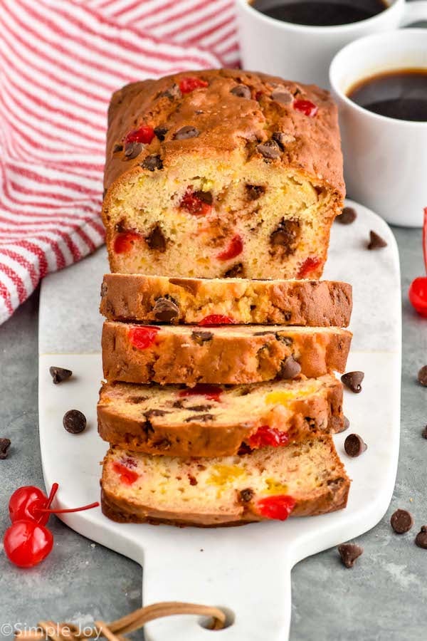 Photo of a loaf of Ambrosia Bread sliced on a cutting board. Two cups of coffee on counter, and cherries and chocolate chips on cutting board beside bread.