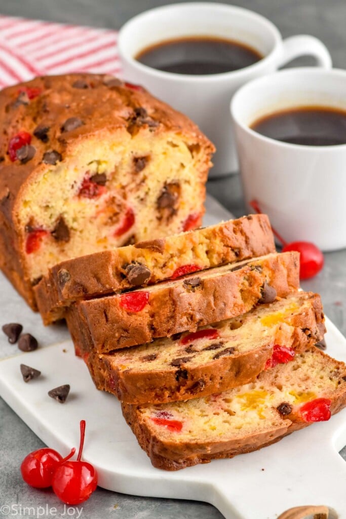 Photo of a loaf of Ambrosia Bread sliced on a cutting board. Two cups of coffee on counter, and cherries and chocolate chips on cutting board beside bread.