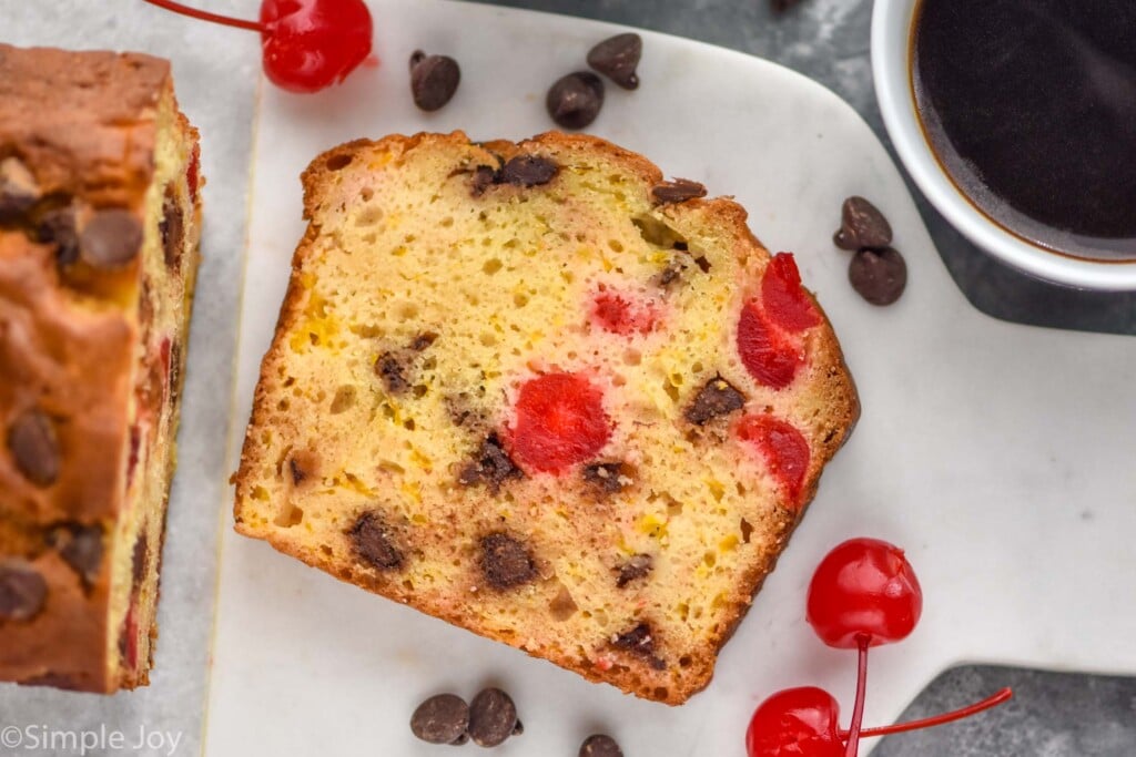 Overhead photo of a slice of Ambrosia Bread next to a loaf of Ambrosia Bread. Cherries and chocolate chips lay on cutting board beside slice.