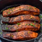 close up of four blackened salmon filets in a cast iron pan garnished with parsley