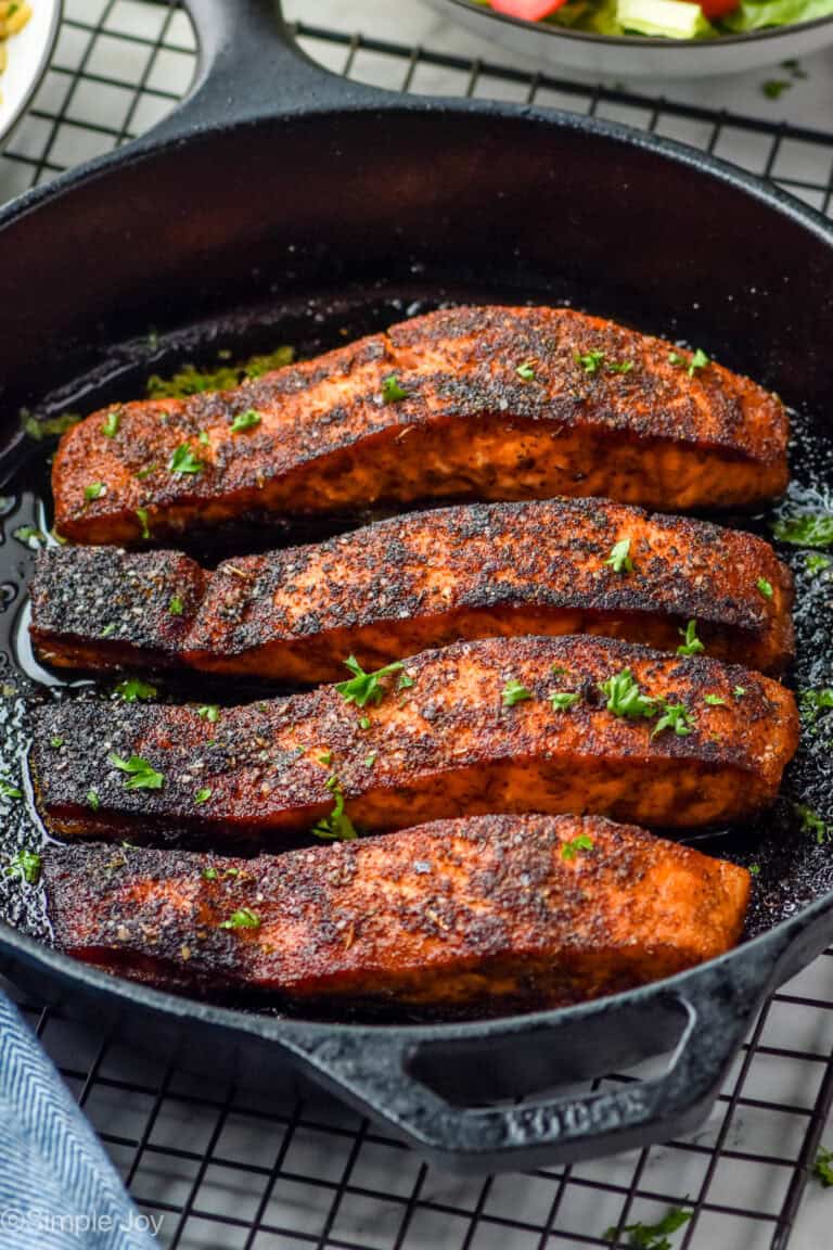 close up of four blackened salmon filets in a cast iron pan garnished with parsley