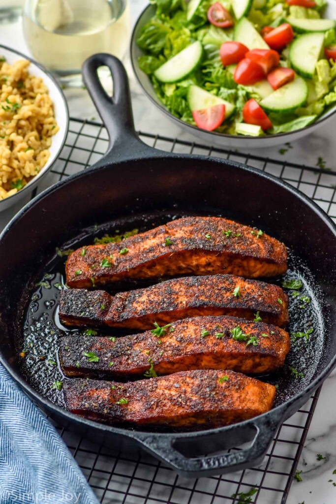 Overhead photo of Blackened Salmon in a skillet. Bowls with salads are beside skillet.