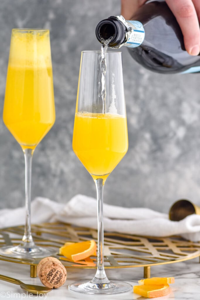 Photo of person's hand pouring champagne into flute glasses of orange juice for mimosa recipe.