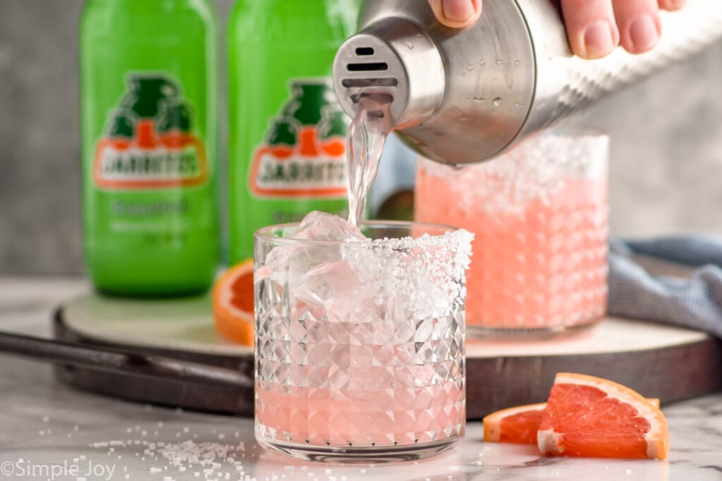 Photo of shaker of ingredients being poured into a tumbler of ice with a salted rim for Paloma recipe. Slices of grapefruit for garnish lay beside glasses. Bottles of grapefruit soda in the background.