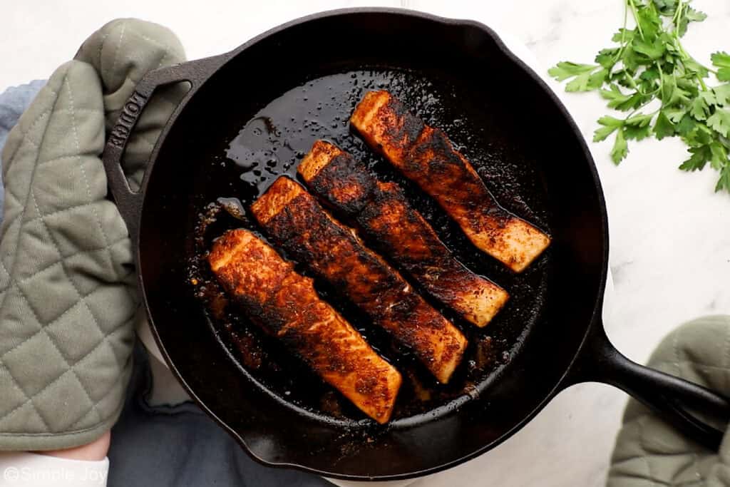 two hands with oven mitts holding a cast iron skillet with blackened salmon filets in it
