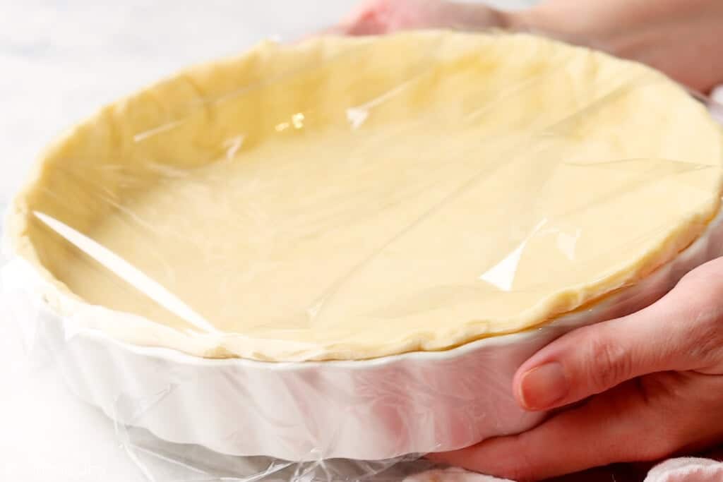 hands holding a pie plate with an unbaked pie crust covered in plastic wrap