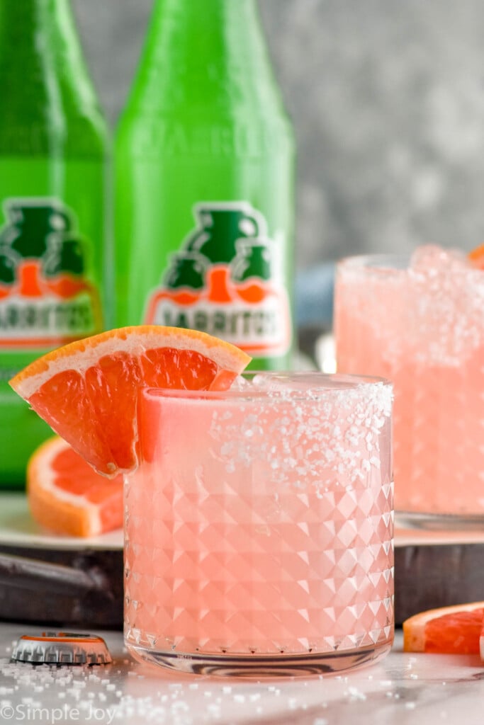 Photo of Paloma cocktails garnished with slice of grapefruit. Bottles of grapefruit soda in the background.