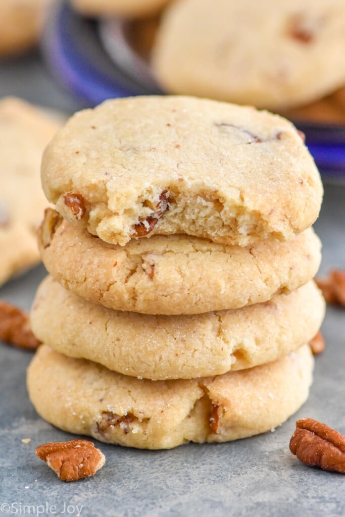 Close up photo of a stack of Pecan Sandies with a bite missing out of the top cookie. Pecans on counter beside stack of Pecan Sandies.