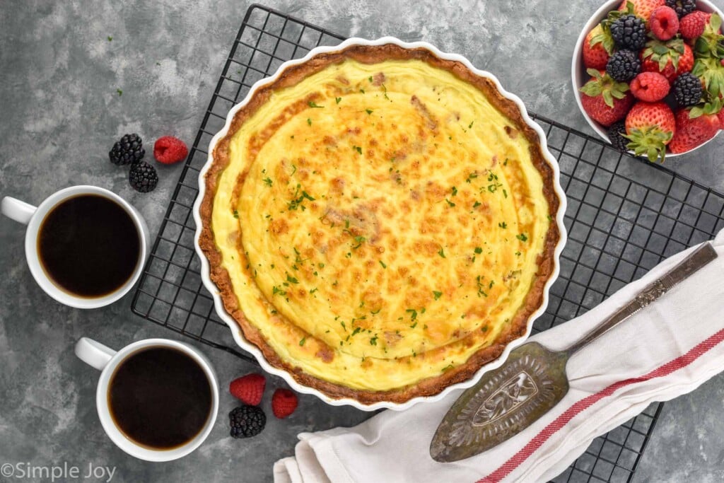 overhead of quiche Lorraine in a fluted pie pan on a cooling rack next to two coffees and a bowl of berries