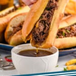 Pinterest graphic for French Dip Sandwich recipe. Image is photo of person's hand dipping French Dip Sandwich into bowl of drippings. Platter of French Dip Sandwiches in the background. Text says, "French Dip simplejoy.com"