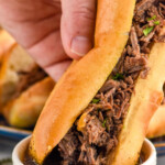 Pinterest graphic for French Dip Sandwich recipe. Text says, "slow cooker French Dip Sandwiches simplejoy.com." Image is photo of person's hand dipping French Dip Sandwich into bowl of drippings.