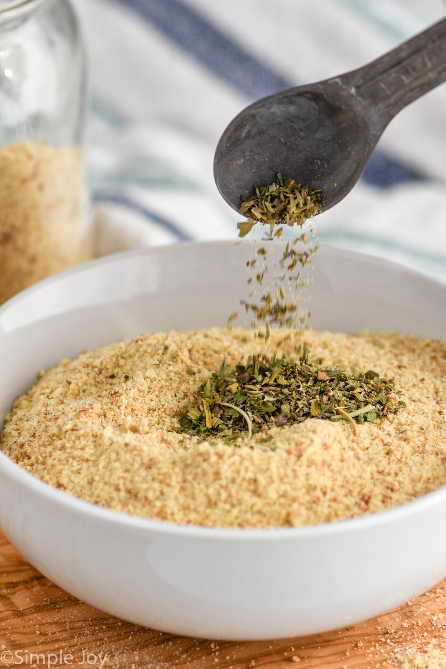 Photo of a bowl of Breadcrumbs with a measuring spoon dumping seasoning into bowl of Breadcrumbs.