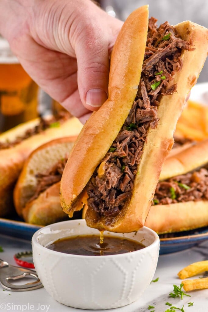 Photo of person's hand dipping French Dip Sandwich into bowl of drippings. Platter of French Dip Sandwiches in the background.