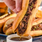 Photo of person's hand dipping French Dip Sandwich into a bowl of drippings. Platter of French Dip Sandwiches in the background.