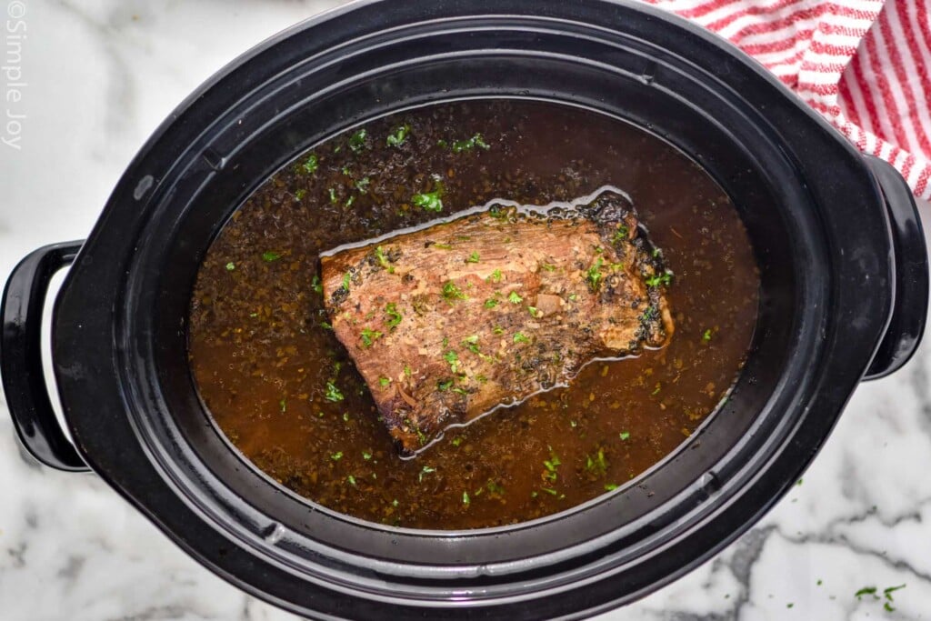 Overhead photo of beef roast and other ingredients cooking in a crockpot for French Dip Sandwich recipe.