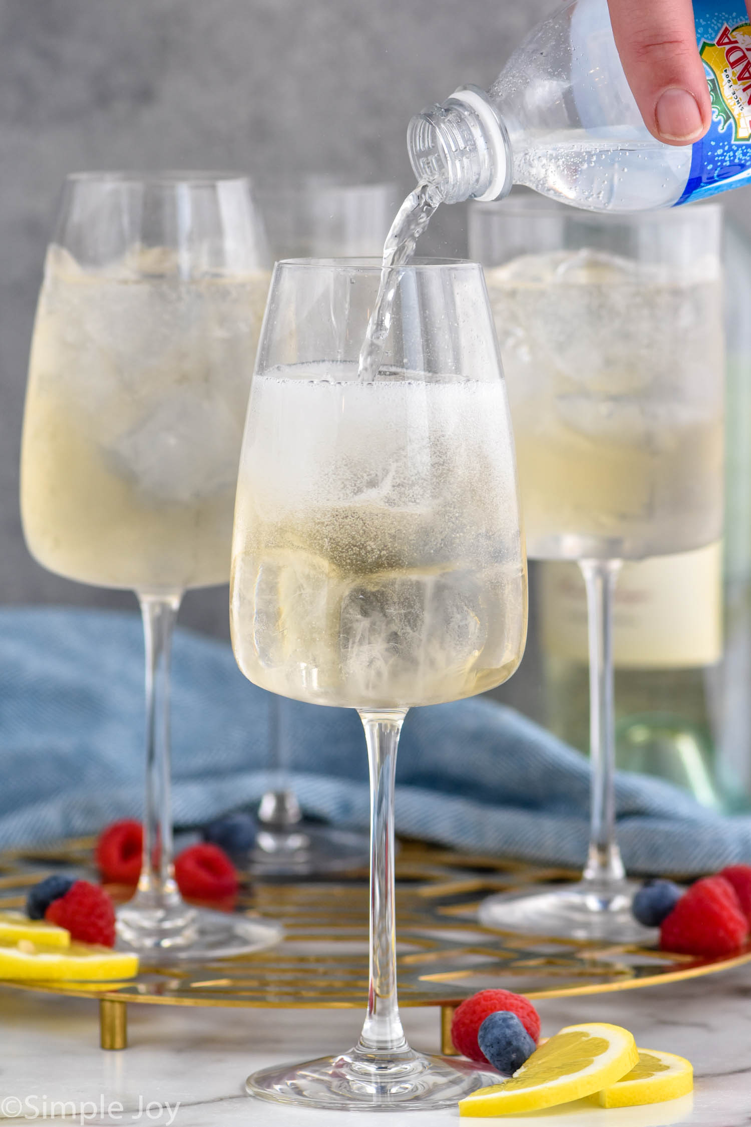Photo of person's hand pouring club soda into a glass of ingredients for White Wine Spritzers. Lemon wedges and berries on counter for garnish.