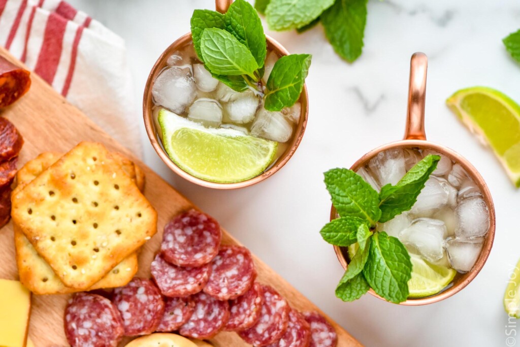 Overhead photo of Kentucky Mules served in copper mugs garnished with lime slices and mint leaves, sitting on counter next to charcuterie board.