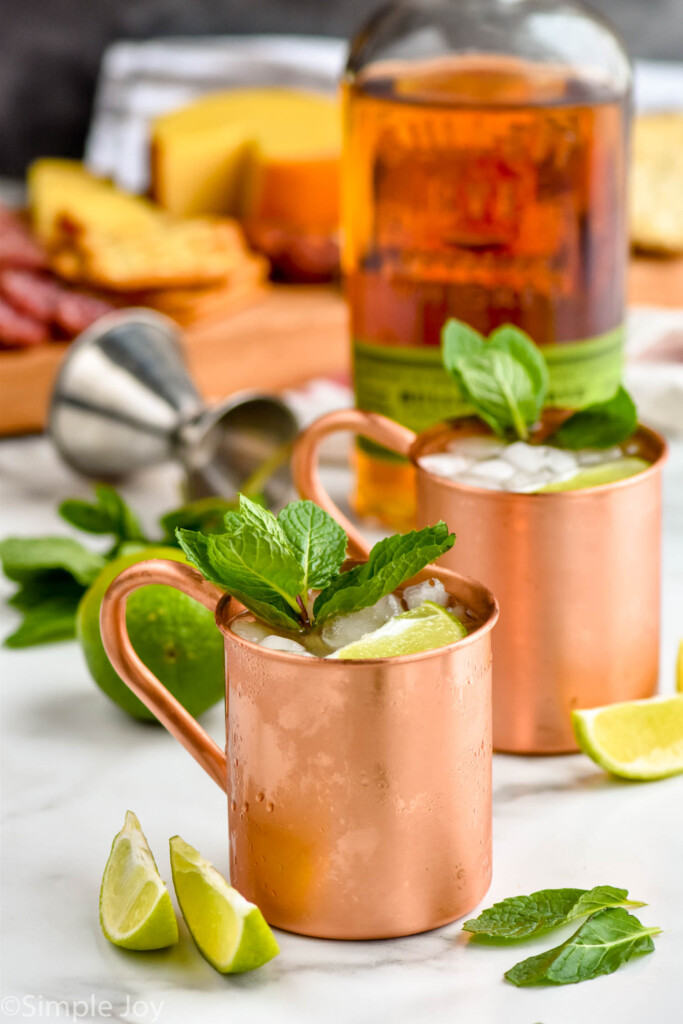Photo of Kentucky Mules served in copy mugs and garnished with lime slices and mint leaves. Bottle of whiskey and a charcuterie board in the background.