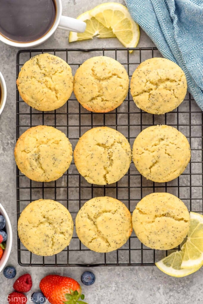 Overhead photo of Lemon Poppy Seed Muffins on a cooling rack. Cup of coffee, slices of lemon, and berries on counter beside rack.