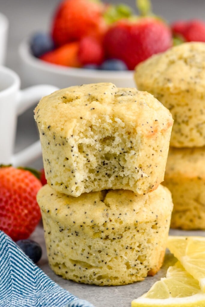 Close up photo of a stack of two Lemon Poppy Seed Muffins with a bite taken out of the top muffin. Bowl of berries in the background.