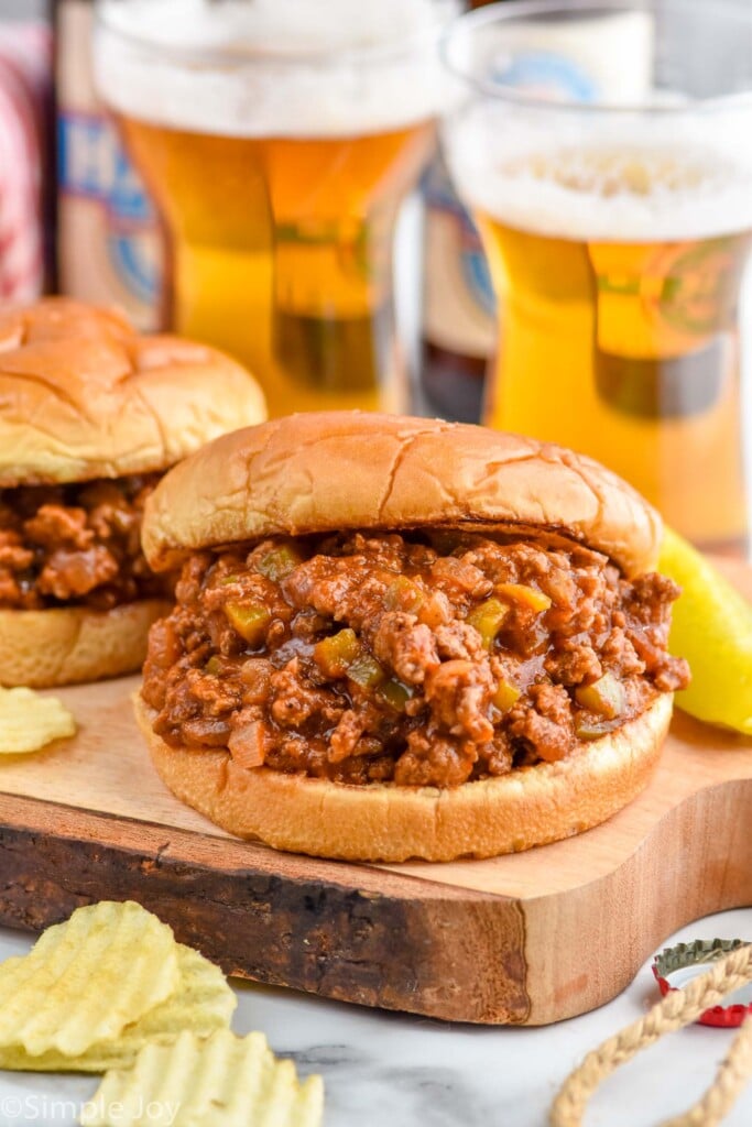 Photo of two Homemade Sloppy Joes with chips, glasses of beer, and pickle spear in the background.