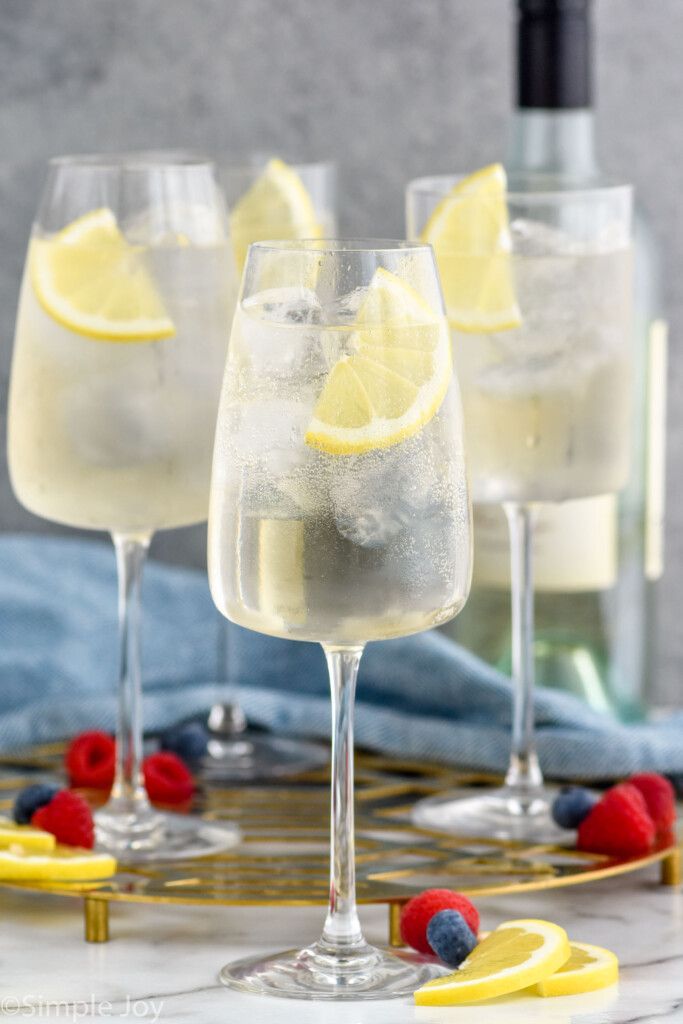 Photo of White Wine Spritzers garnished with lemon wedges. Berries for garnish on counter beside wine glasses.