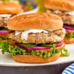 Photo of Greek Turkey Burgers served on a bun with yogurt sauce, lettuce, tomato, pickles, and onions.